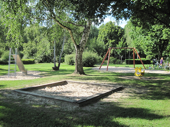Blick auf den Sandkasten, die Rutsche und rechts die Schaukel und ein gelbes Wipptier auf dem Spielplatz im Park am Entenweiher