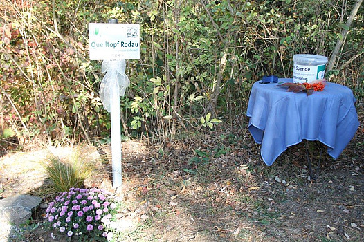 Festlich geschmücktes Schild der Rodauquelle mit Spendeneimer auf einem Tischchen rechts davon.