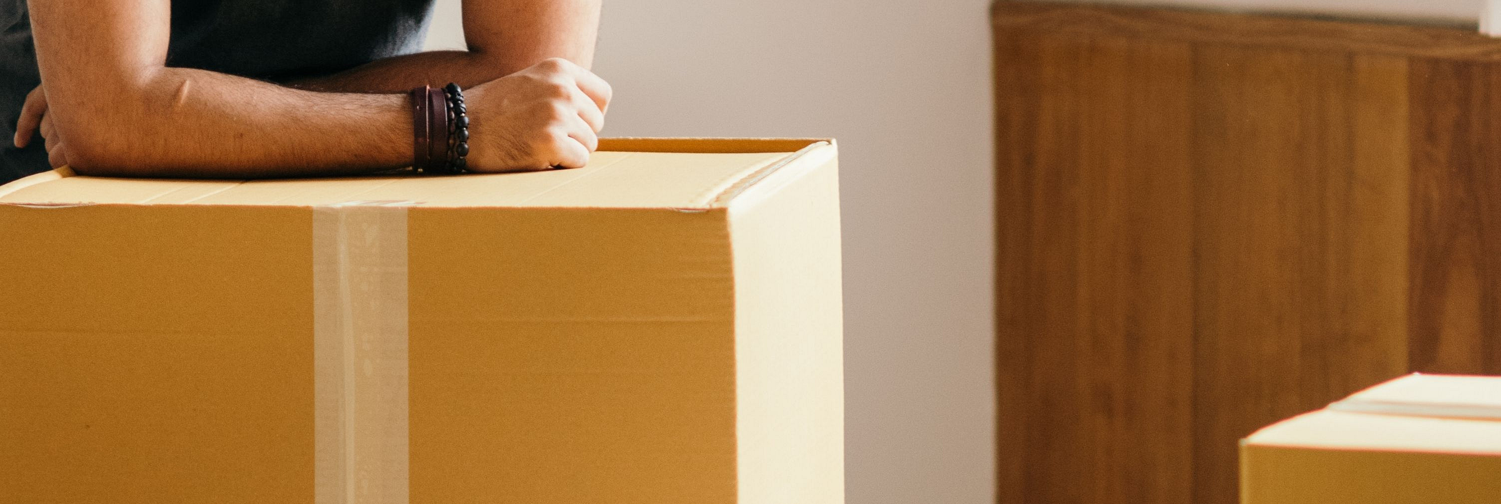 Oberkörper einer Person mit verschränkten Armen auf einem großen Karton in einem Raum mit weiteren Kartons und Holzwänden